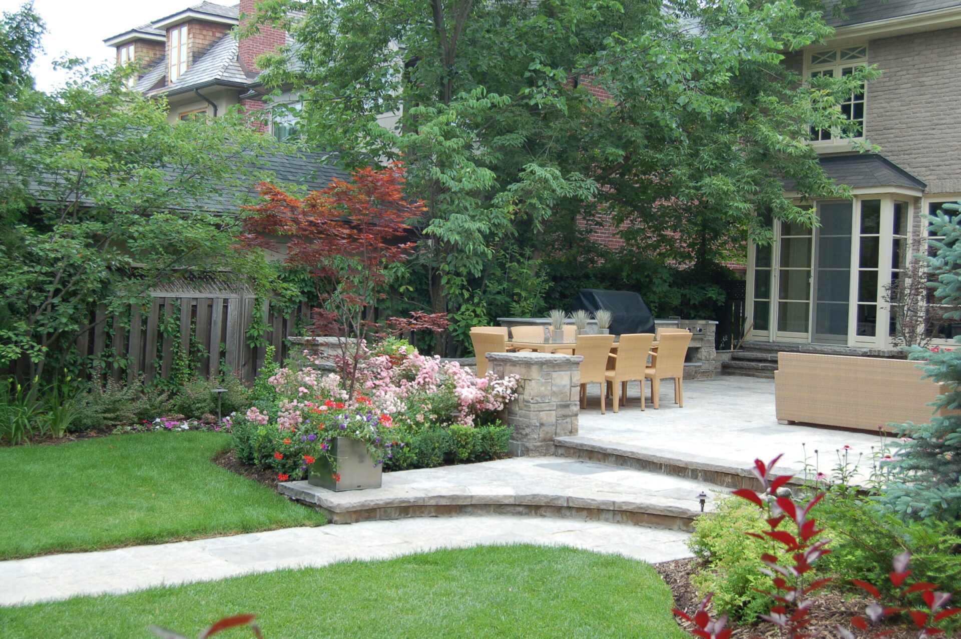 A serene garden with lush greenery, a patio featuring a table, chairs, and a barbecue grill, adjacent to a brick house.