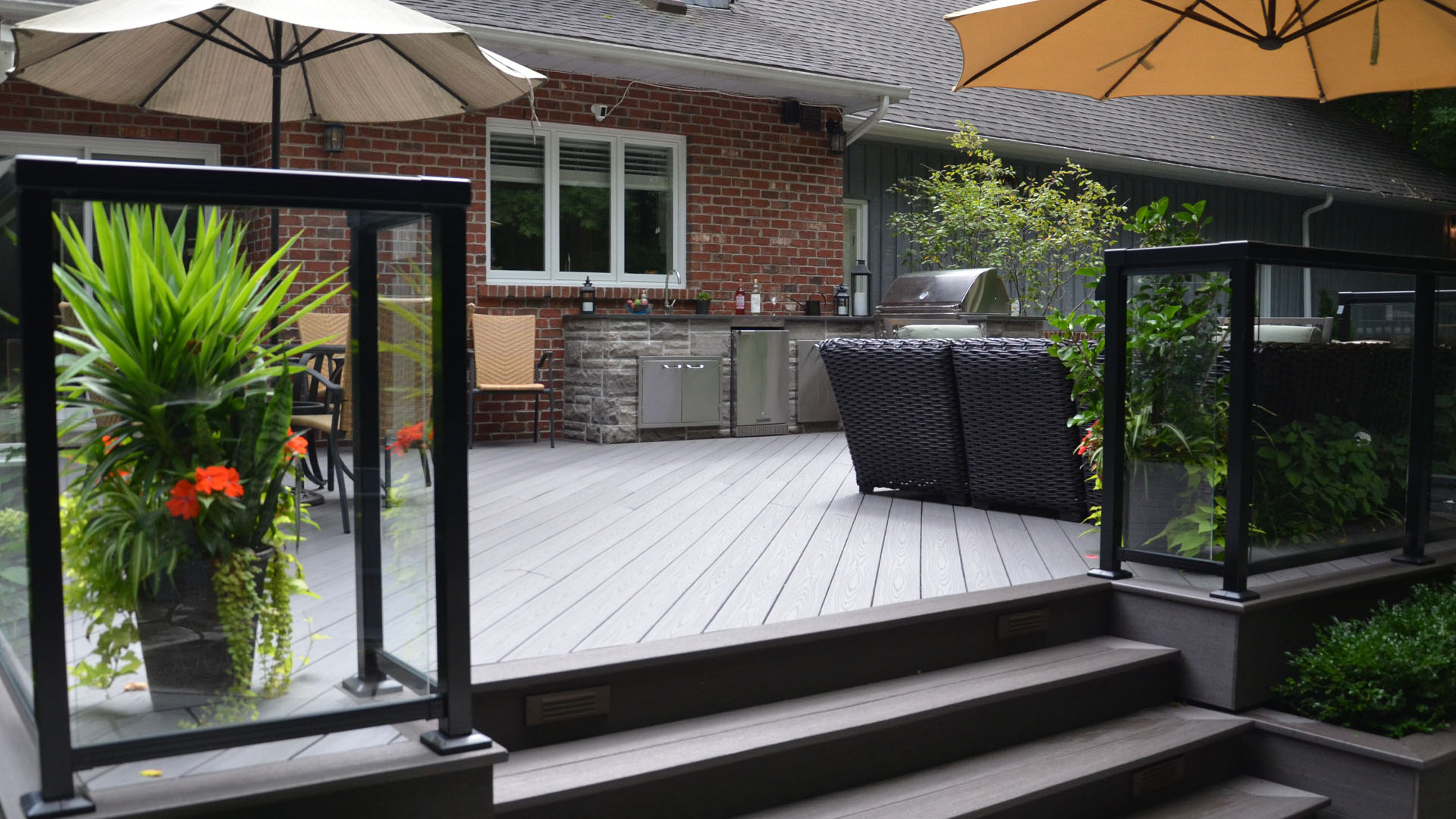 A modern outdoor patio with gray decking, glass railings, potted plants, wicker furniture, and large umbrellas next to a brick house and grill.