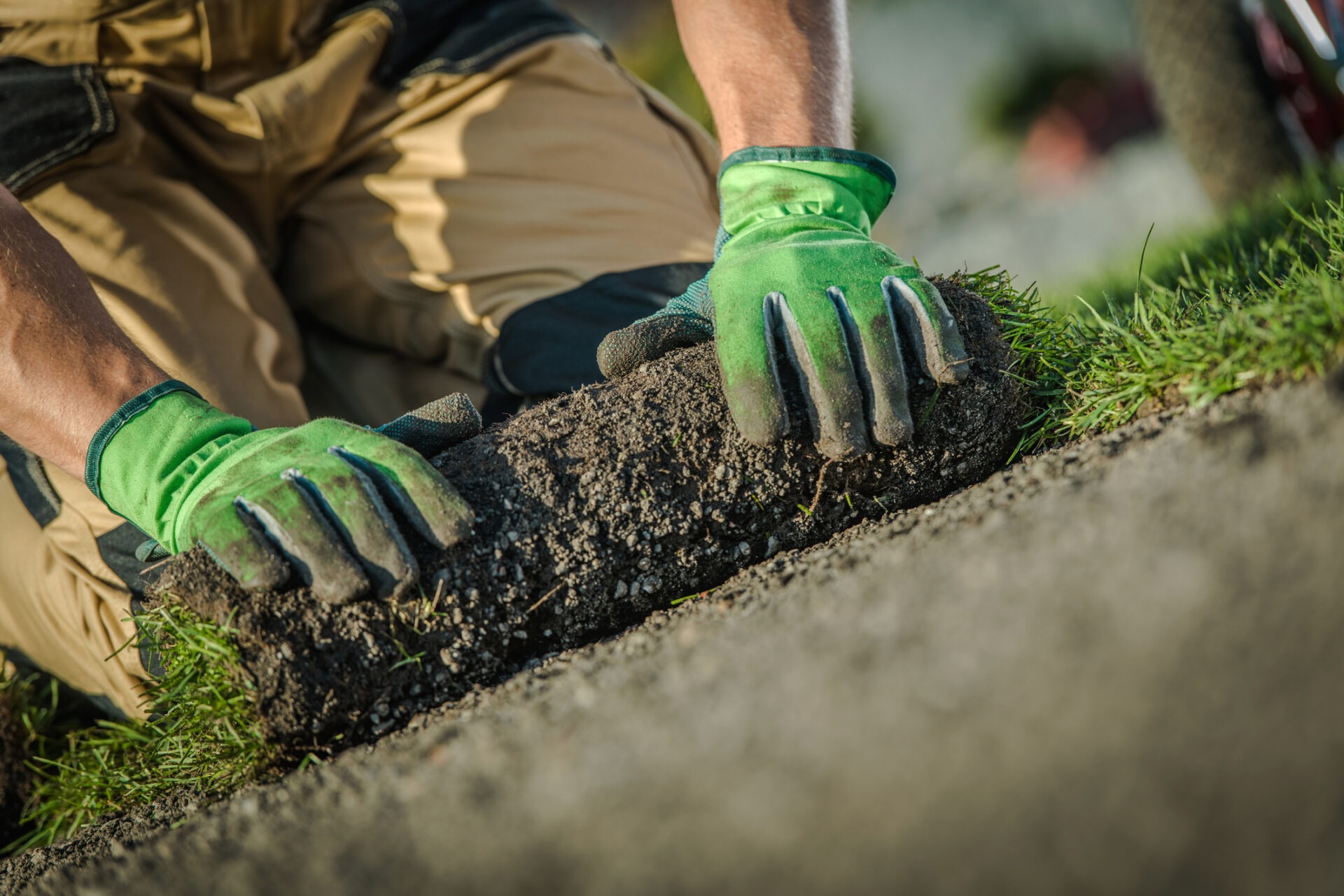 A person in green gloves is laying grass sod on soil, focusing on details and alignment in outdoor landscape work.