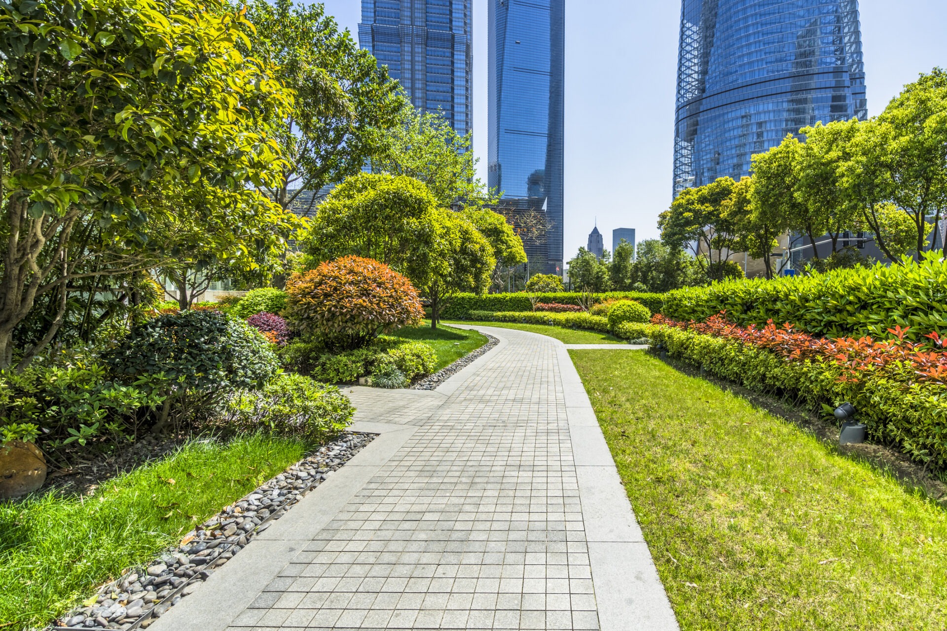 Path winding through a vibrant garden with skyscrapers in the background, possibly in a modern urban park setting. Lush greenery surrounds the area.