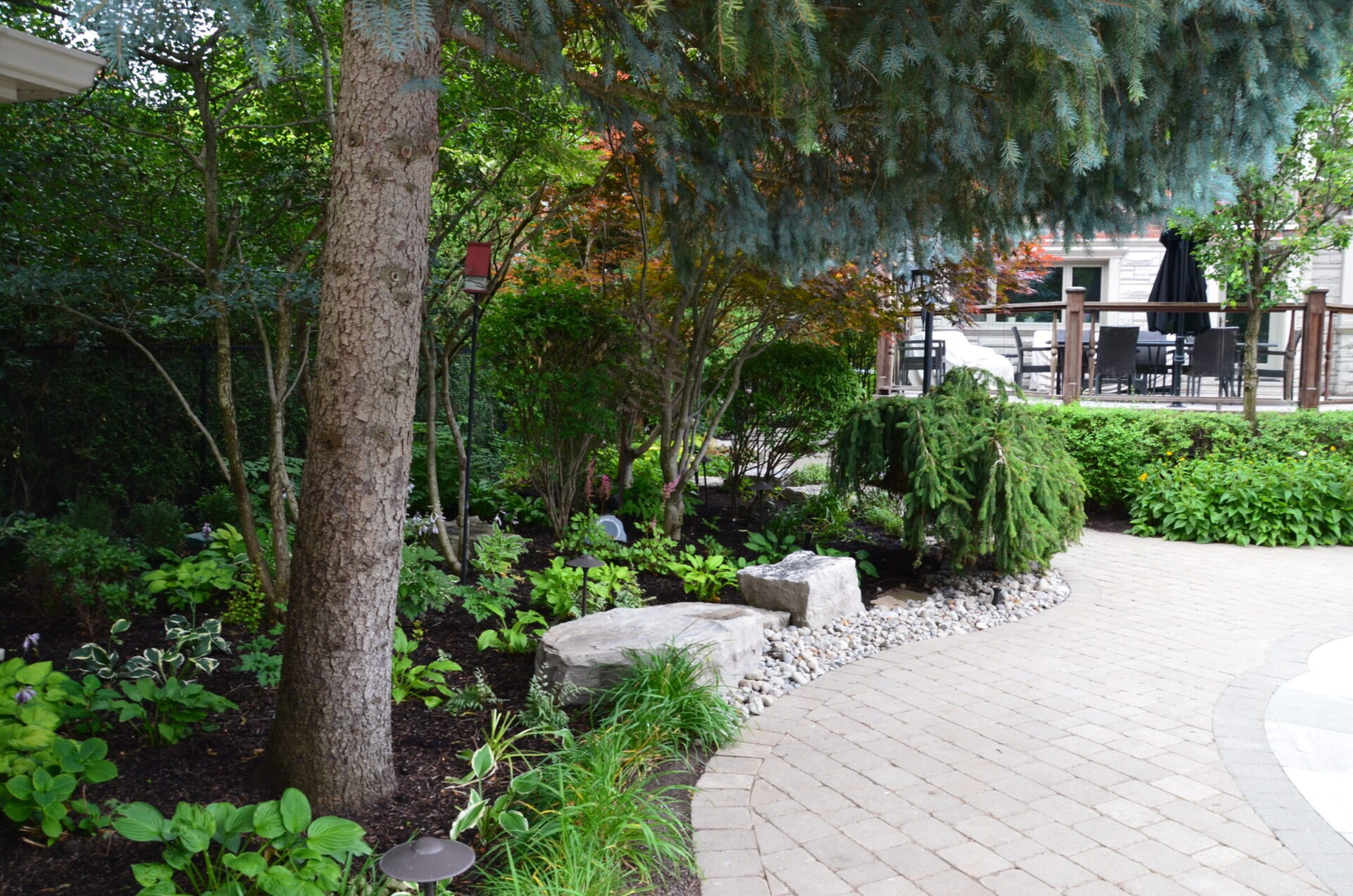 A serene garden with lush greenery, stone path, and surrounding foliage. Patio furniture is visible in the background under umbrellas.