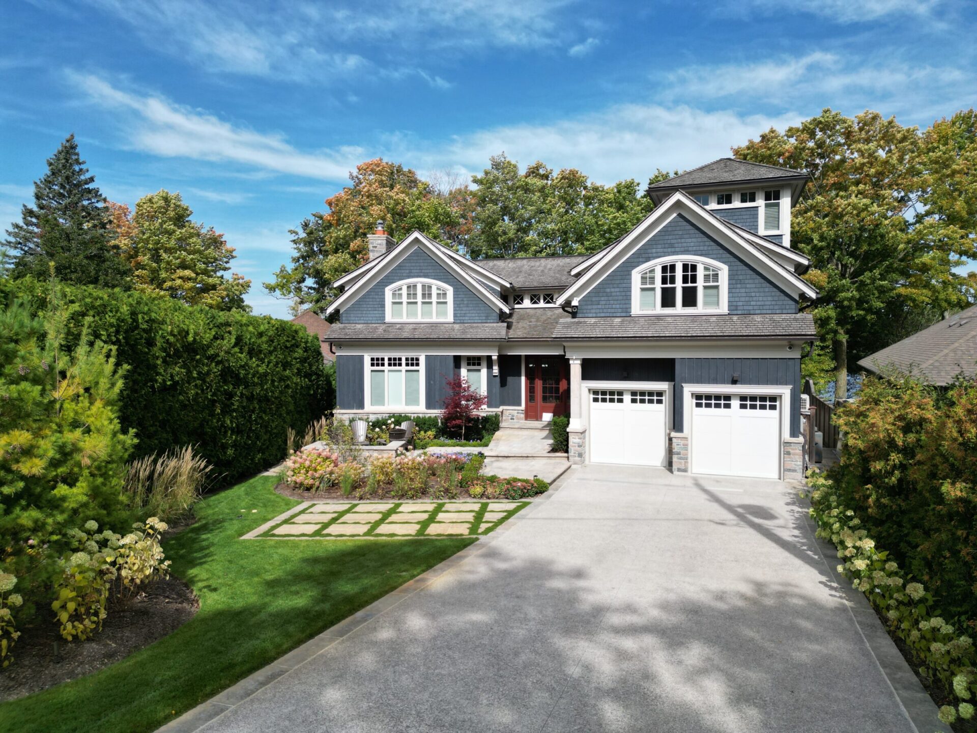 Two-story gray house with white trim, surrounded by trees and a neat garden. Wide driveway in front, under a clear blue sky.