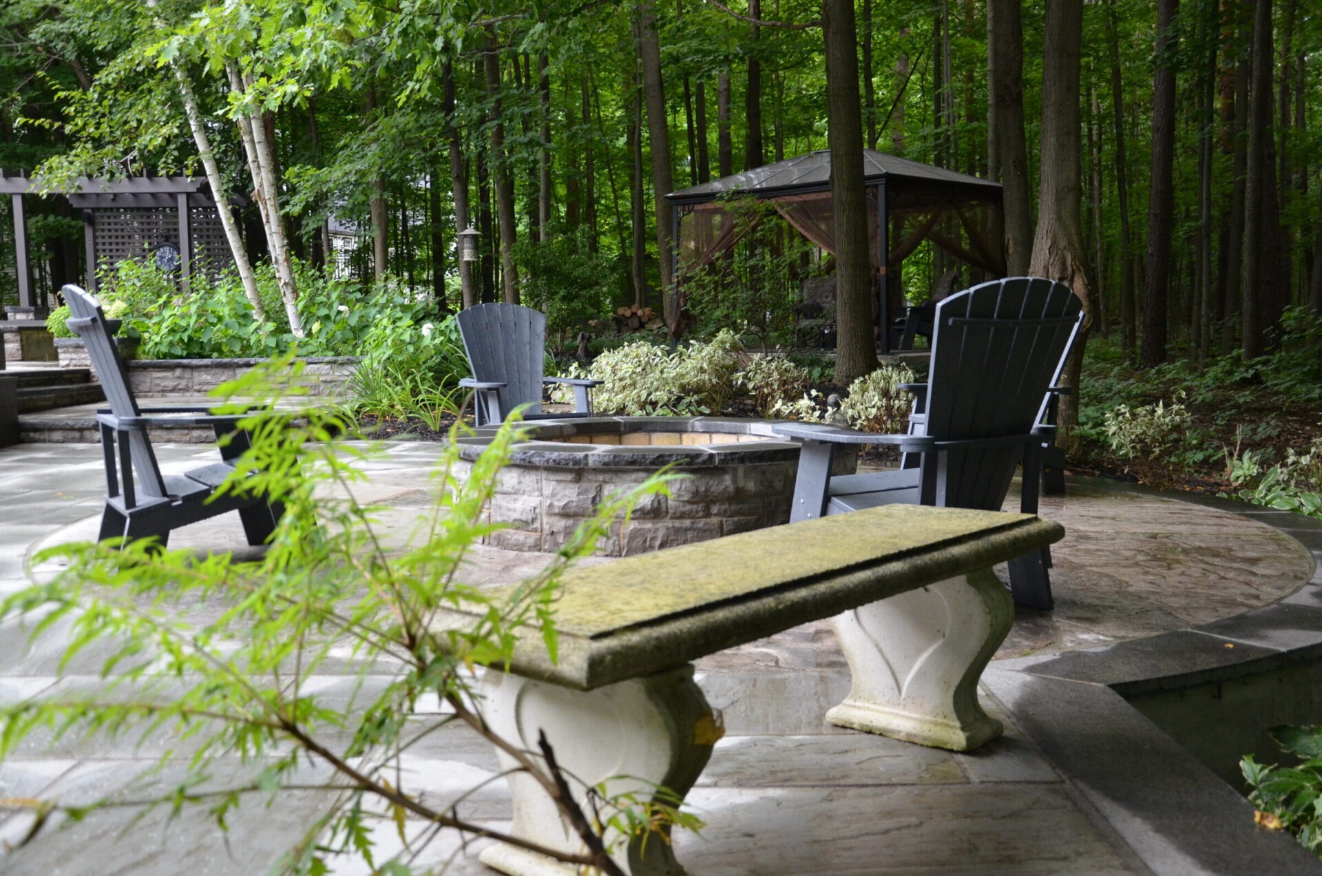 A serene outdoor patio with Adirondack chairs, a fire pit, and a stone bench surrounded by lush green trees and garden plants.