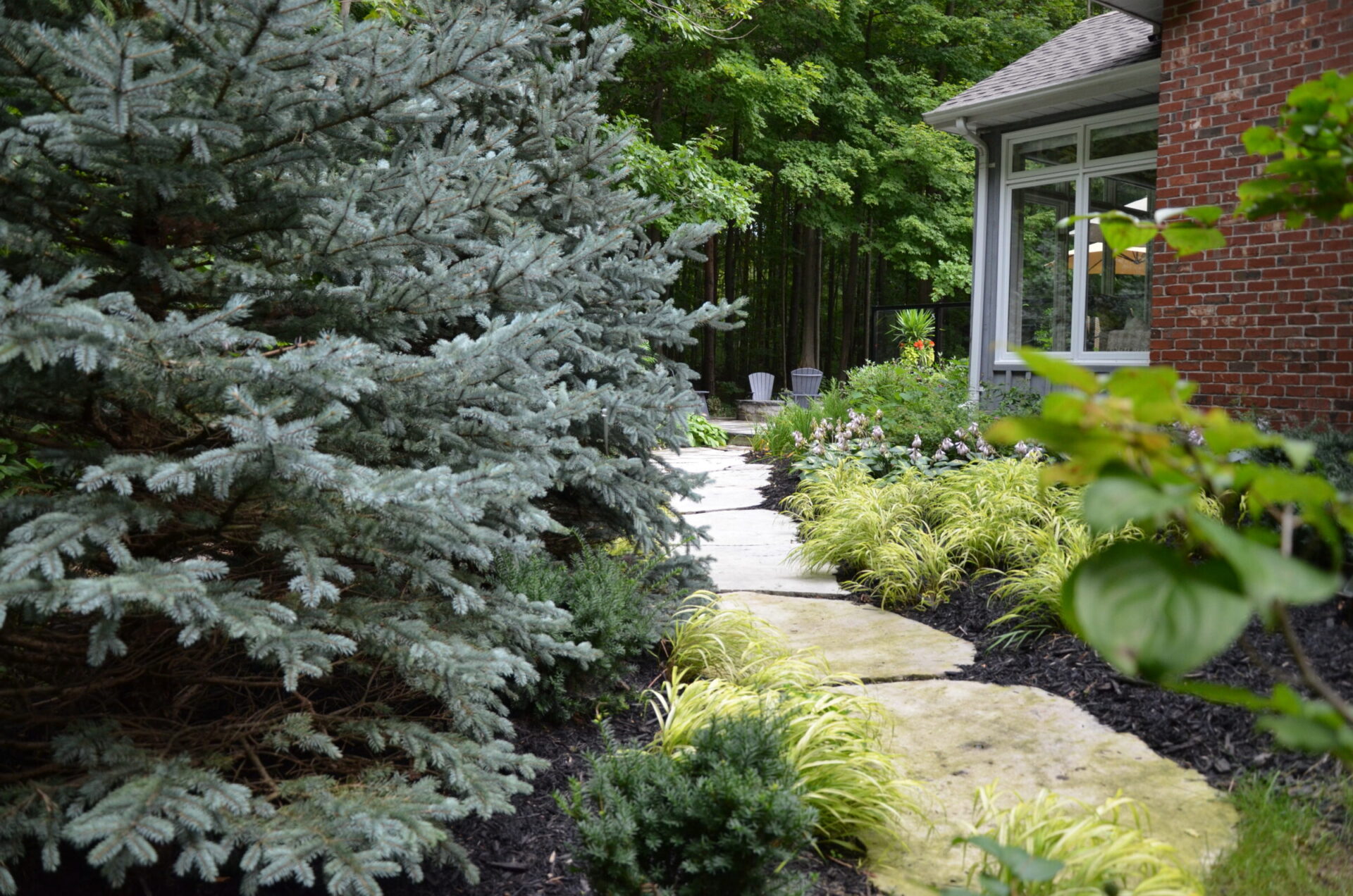 A serene garden scene with a winding stone path, lush greenery, and a brick house in the background, surrounded by dense woods.