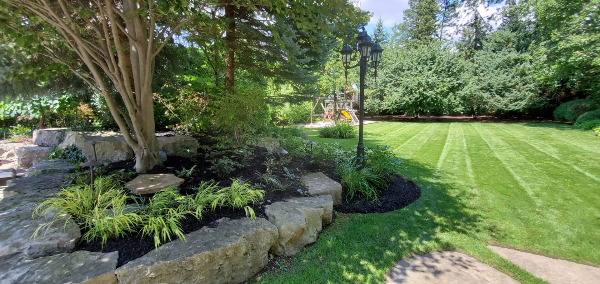 A well-maintained garden features a green lawn, stone-bordered flower beds, a tall lamppost, and a background of trees under a clear sky.