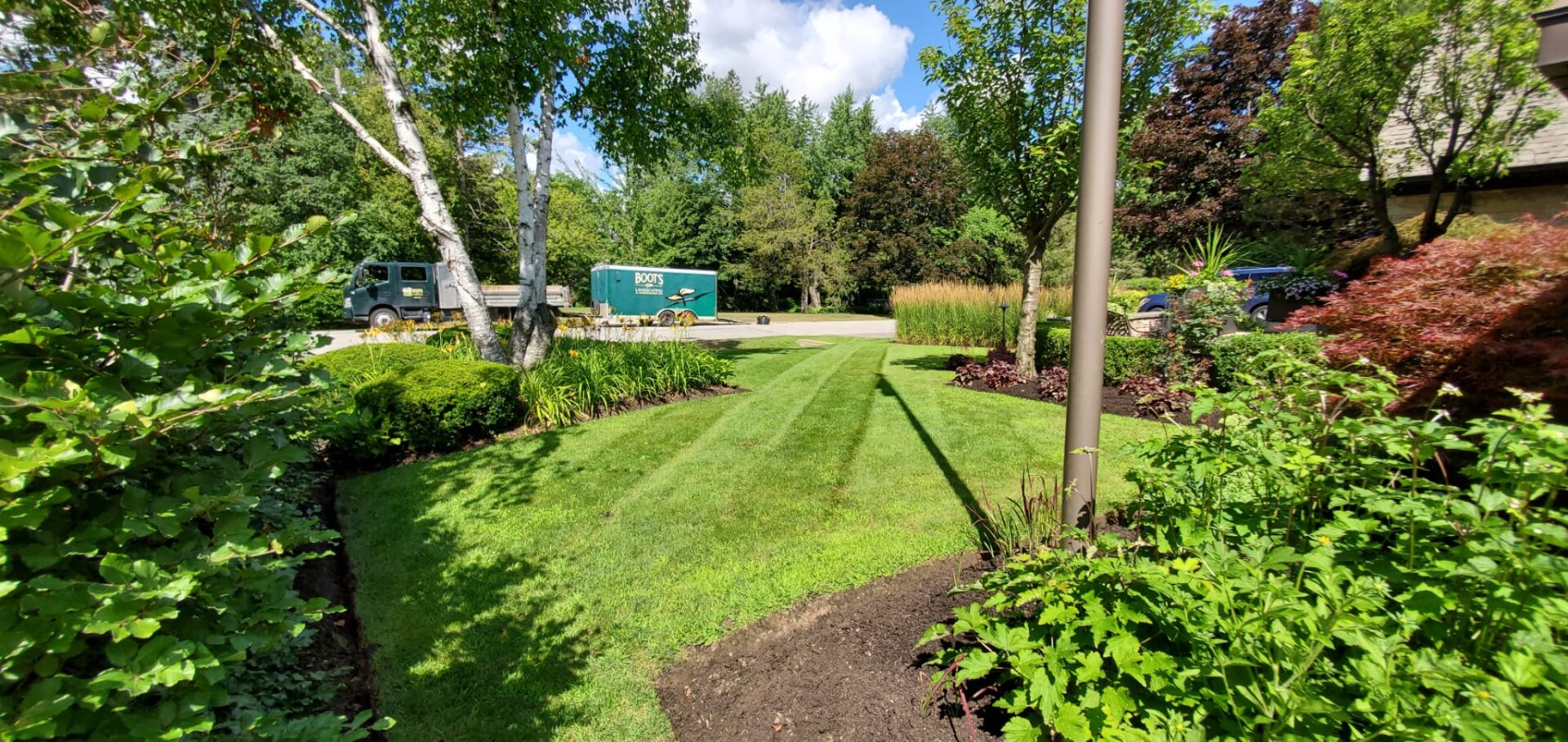 A lush garden with trimmed grass and trees, a parked truck, and trailer in the background under a sunny blue sky.