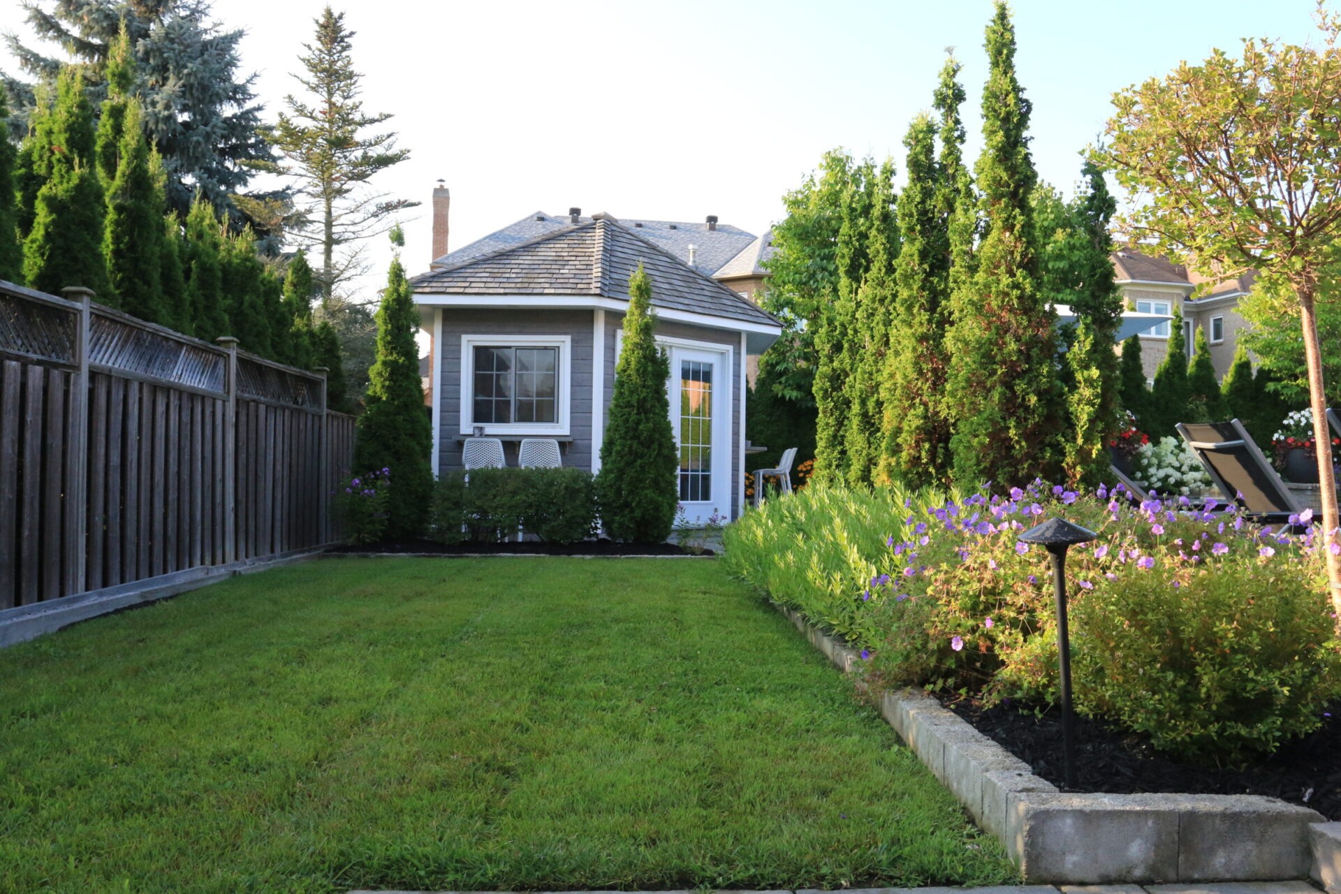 A small backyard with a grassy lawn, surrounded by trees and flowers, leads to a cozy, white garden shed with a peaked roof.