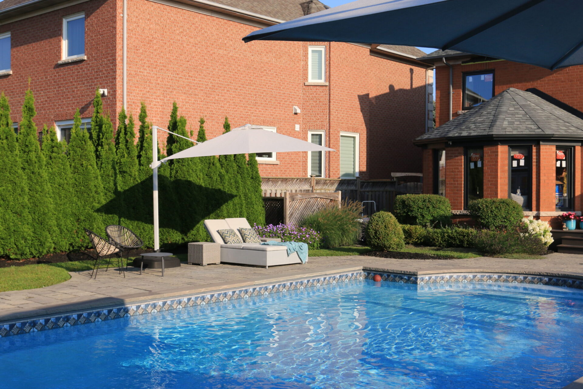 A backyard scene with a swimming pool, patio seating, umbrella, and well-maintained garden by a red brick house under a clear blue sky.