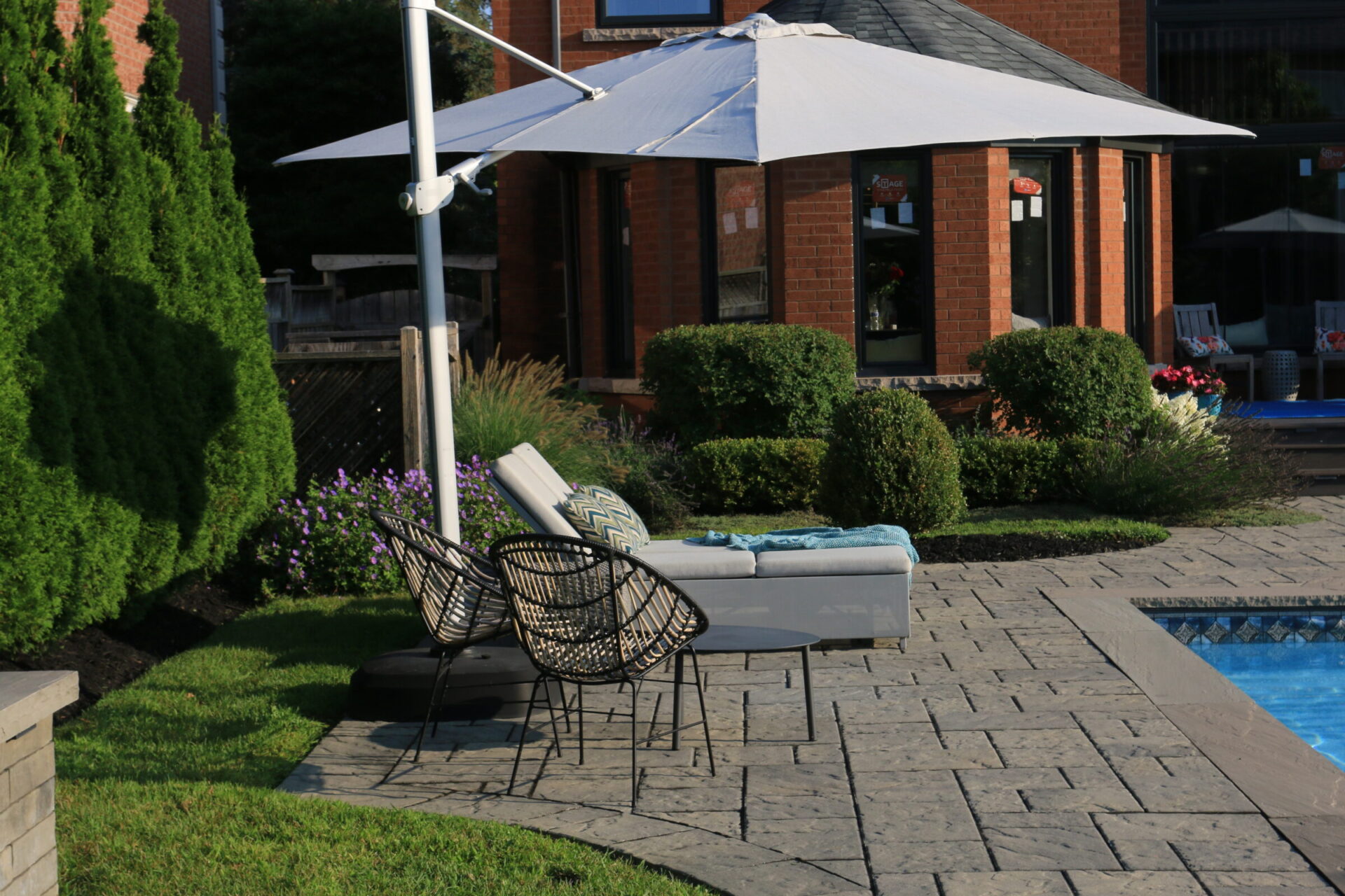 A backyard patio with lounge chairs, umbrella, and a pool. Brick house in the background, surrounded by greenery and manicured shrubs.