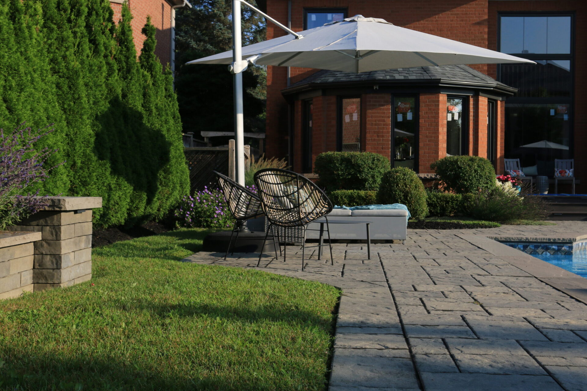 A tranquil backyard scene with patio chairs, large umbrella, and well-maintained greenery beside a pool. Brick house visible in the background.