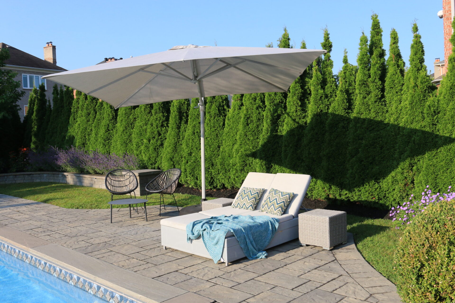 A sunny backyard poolside setup includes lounge chairs, a large umbrella, and lush greenery, creating a private and relaxing outdoor space.