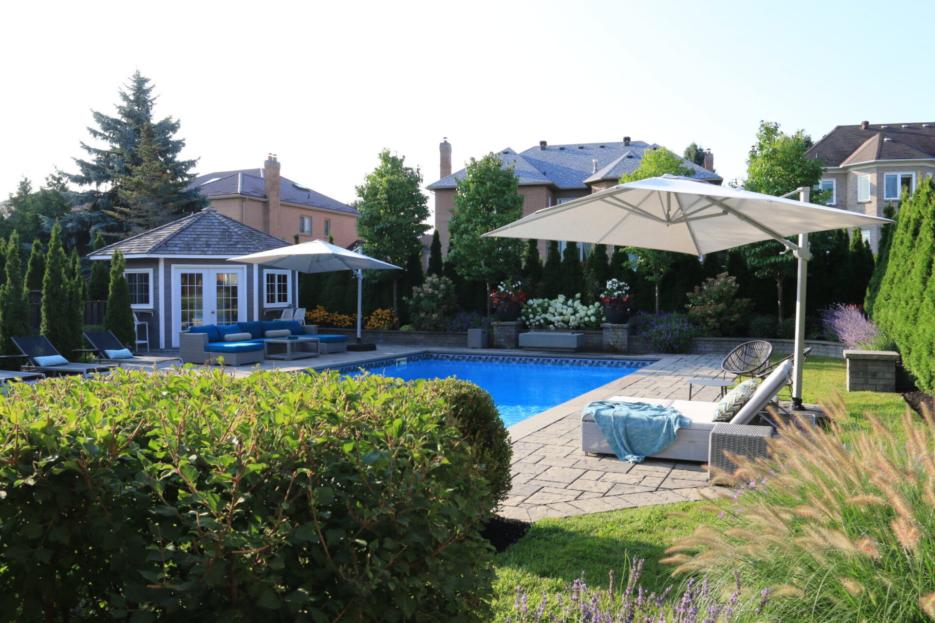 Backyard with a swimming pool, patio furniture, umbrellas, and lush greenery. Neatly landscaped with surrounding trees and a small pool house structure.