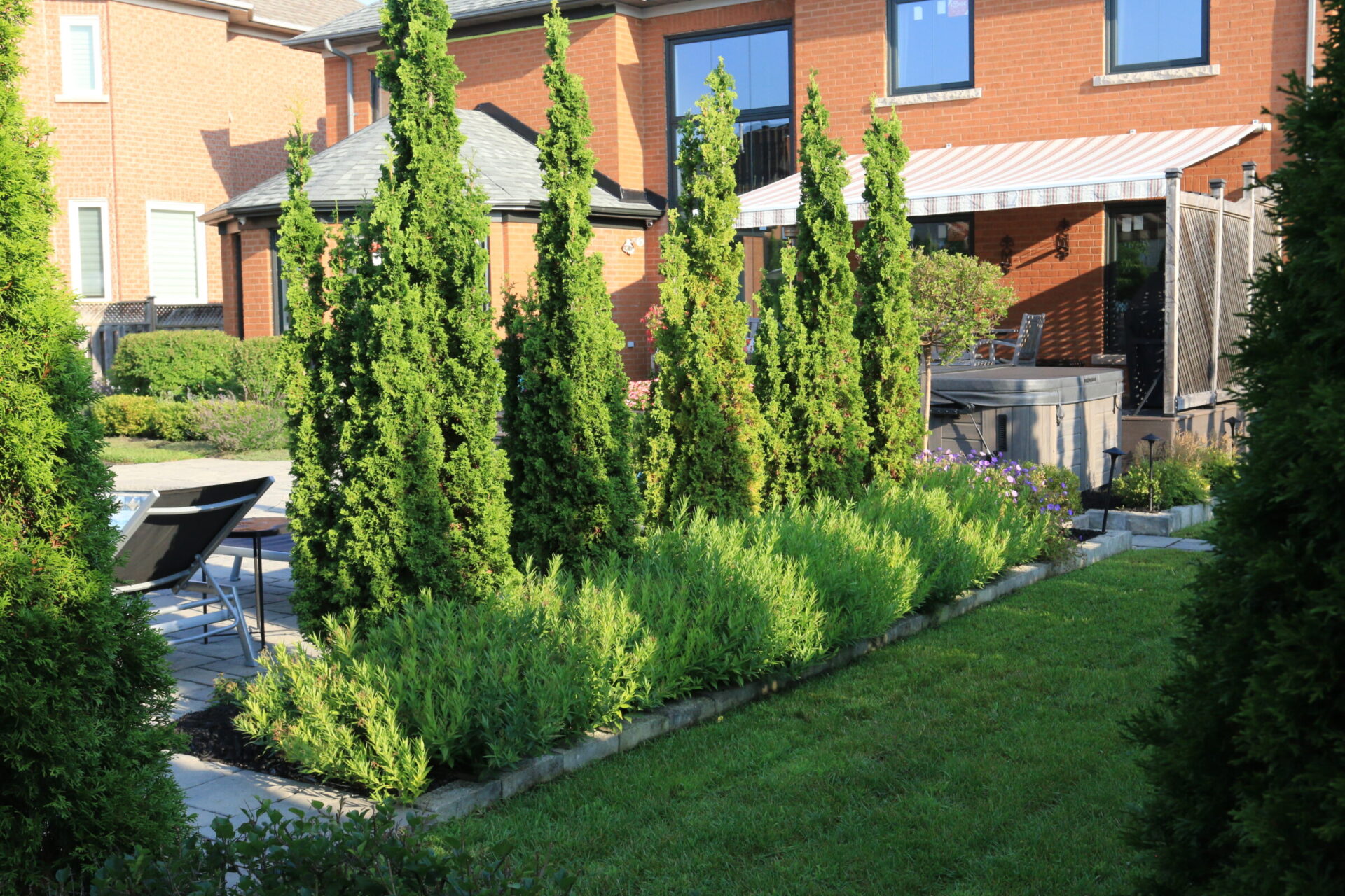 A well-maintained backyard features lush greenery, a small patio, and a red brick house with large windows and a shaded area.