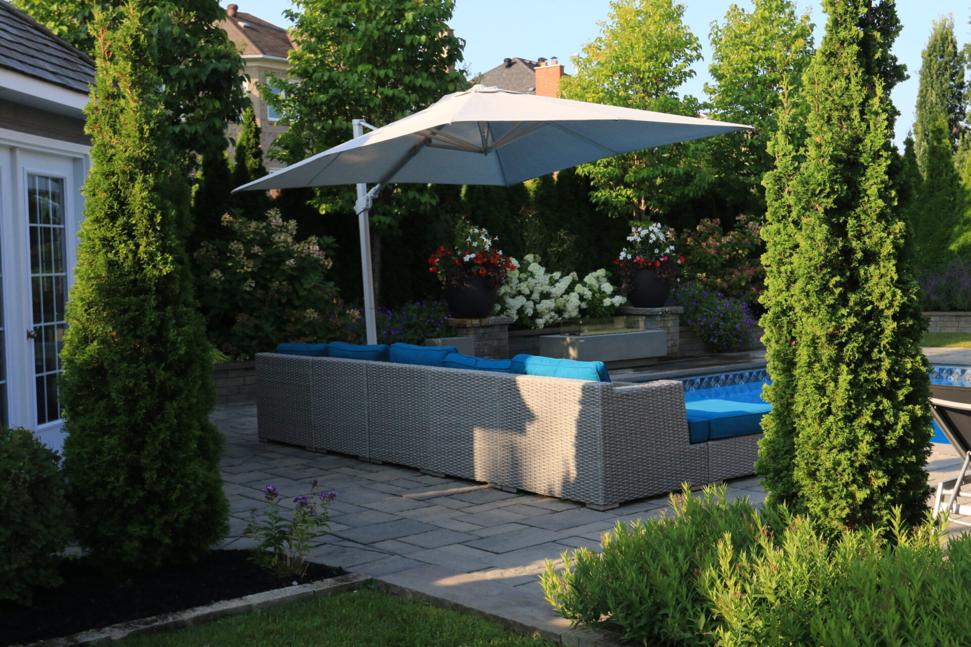 A patio with wicker furniture, blue cushions, and a large umbrella. Surrounded by trees and colorful flowers, creating a relaxing outdoor space.