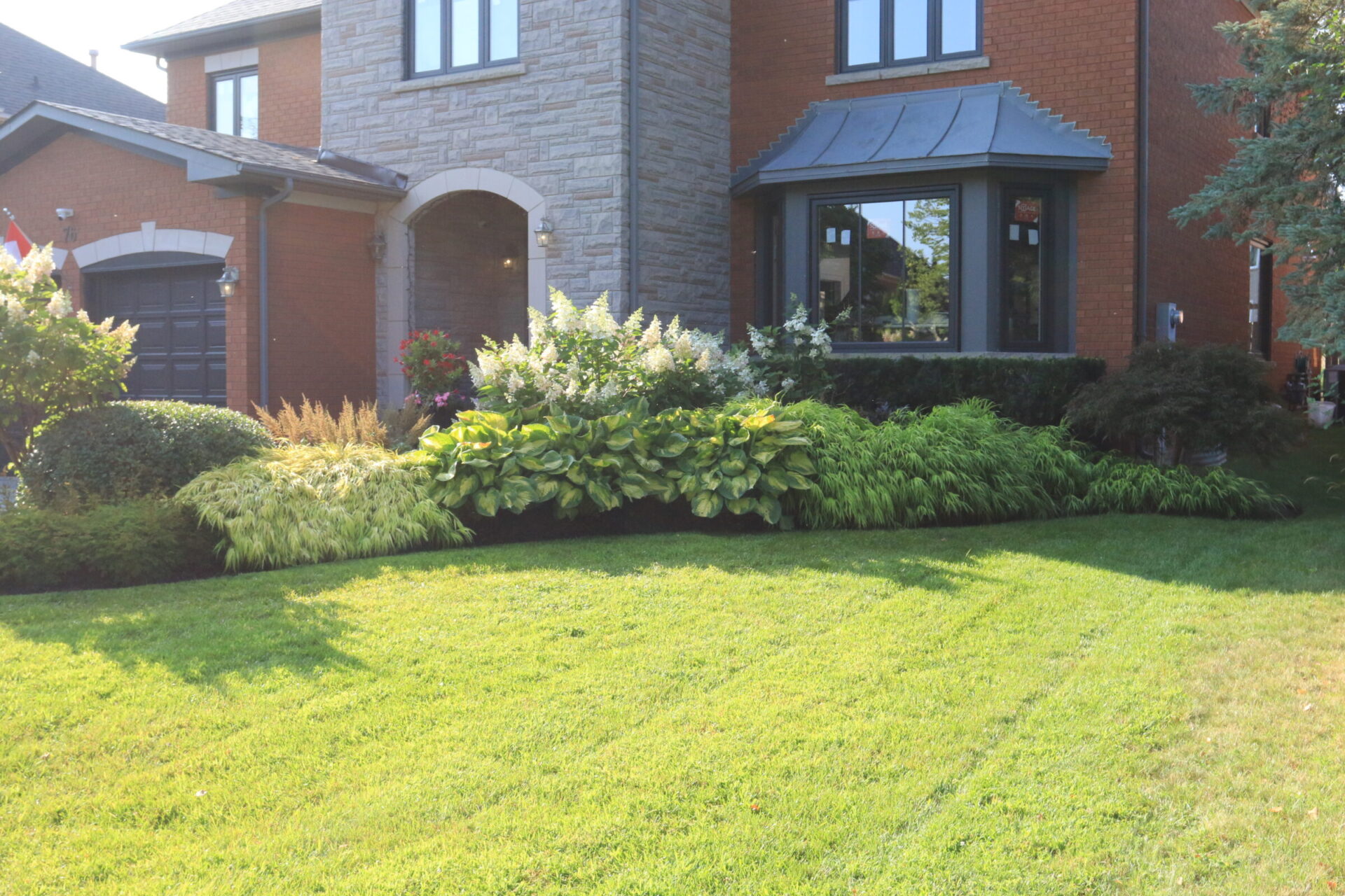 A suburban house with brick and stone facade, featuring well-maintained green lawn, lush shrubs, and a large bay window in front.