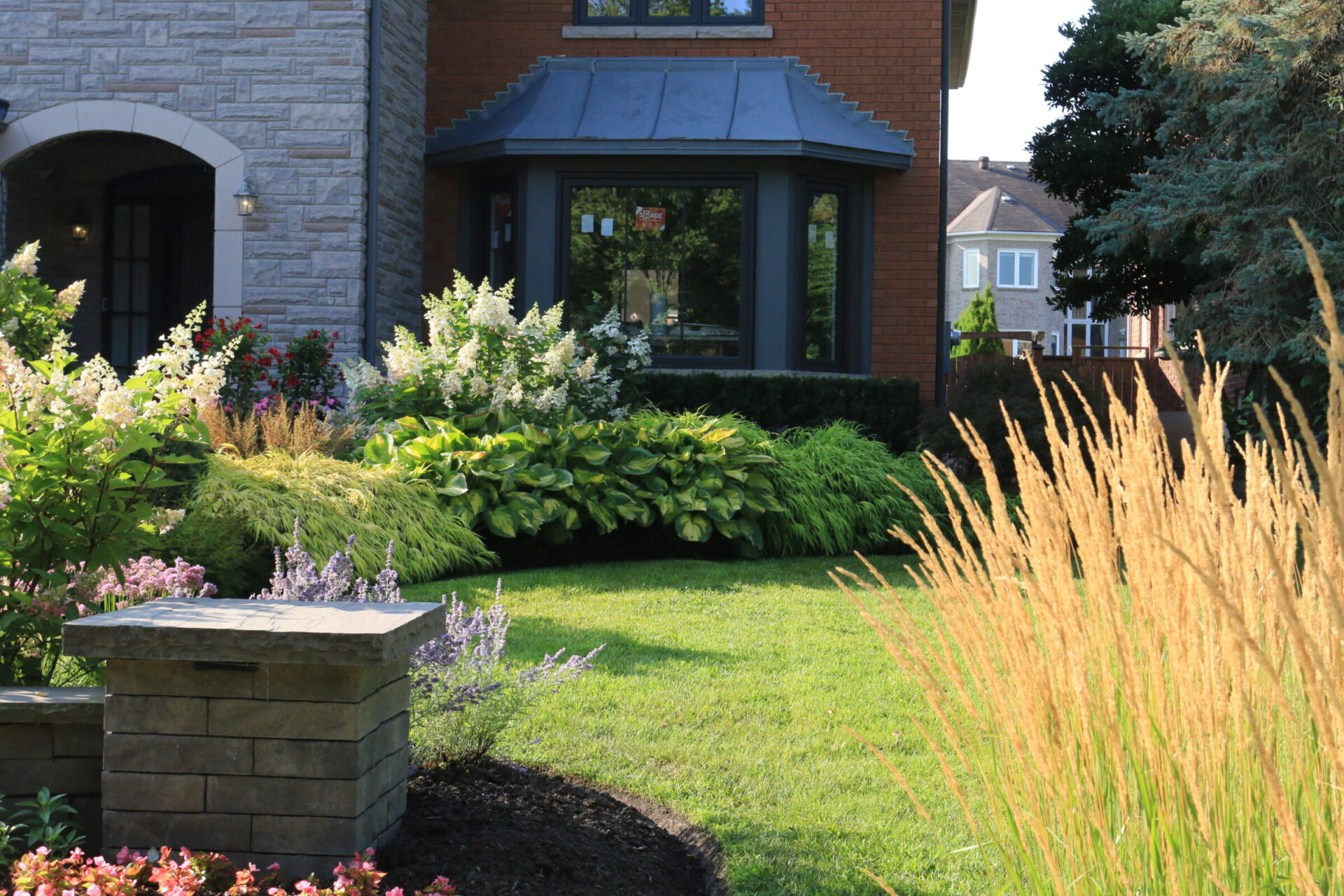 A well-manicured garden with lush greenery and flowers surrounds a brick house with large windows. No identifiable landmarks or historical buildings are visible.