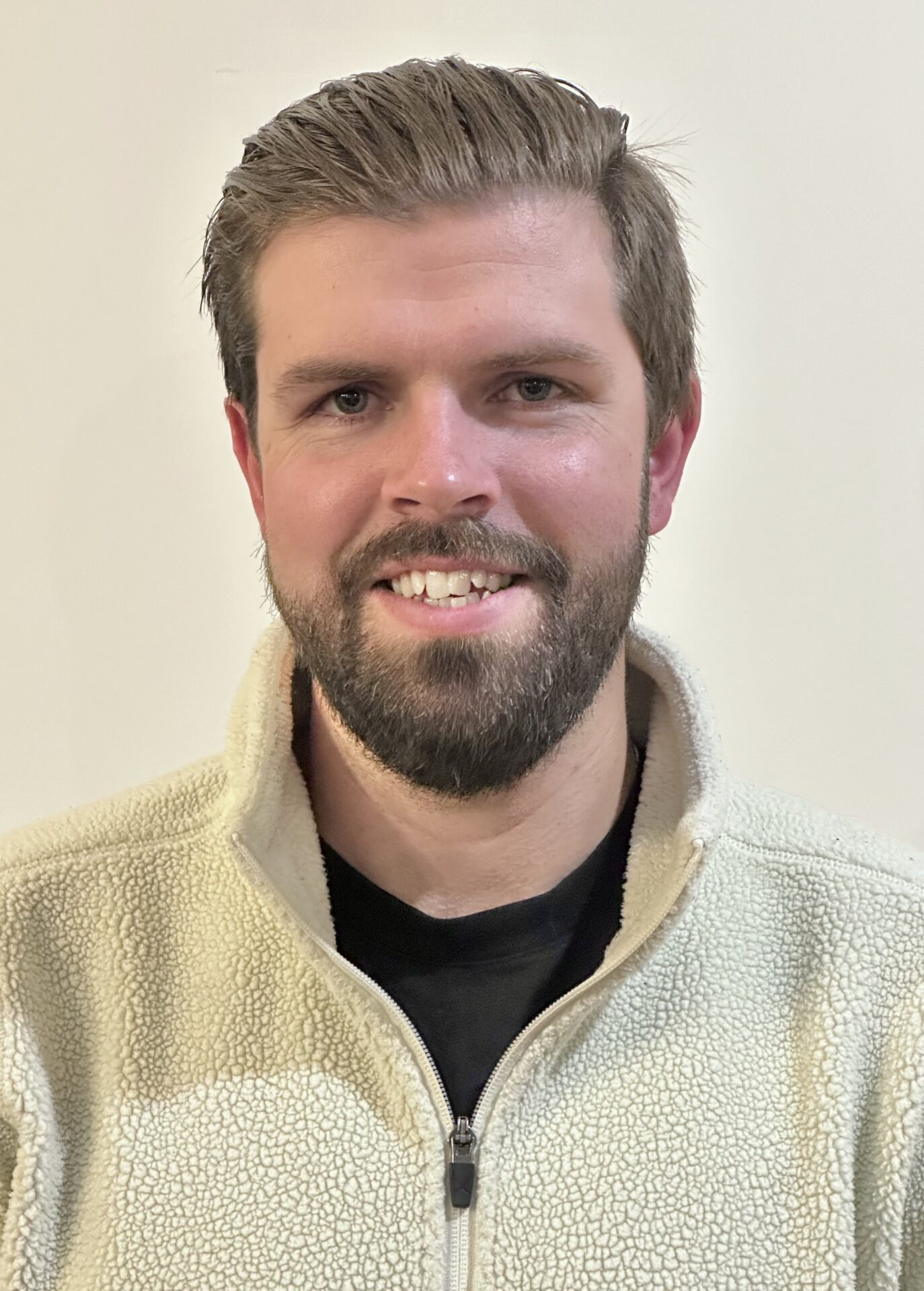 A person with a beard and short hair is smiling at the camera, wearing a light-colored fleece jacket against a plain background.