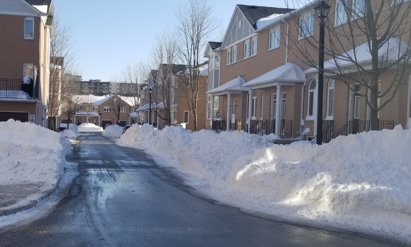 A snowy residential street lined with row houses, clear blue sky overhead, and snow piles along the sidewalks in a suburban neighborhood.